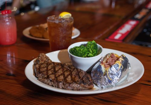 A plate with a grilled steak, baked potato with toppings, steamed broccoli, and a drink in the background.