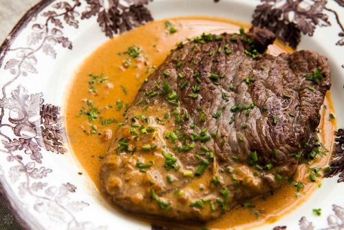A piece of beef steak in a creamy sauce, garnished with chopped herbs, served on a decorative plate.