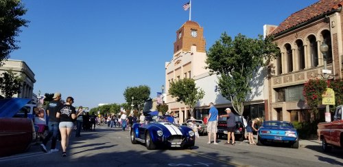 A vibrant street scene with classic cars, people walking, and a clock tower in the background under a clear blue sky.