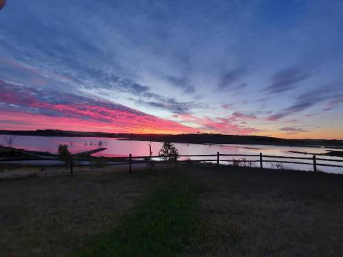 A serene landscape at sunset, featuring vibrant pink and purple clouds over a calm lake, with a wooden fence in the foreground.