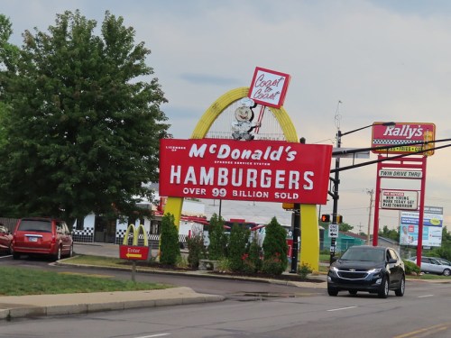 A McDonald's sign advertising "HAMBURGERS" with a large number sold, next to a tree and other businesses.