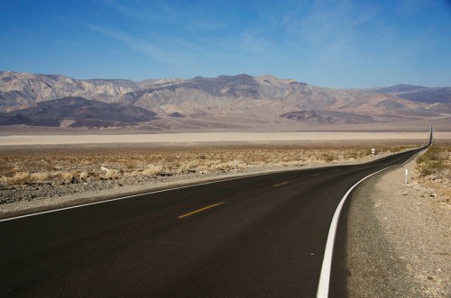 A winding road stretches through a vast desert landscape with mountains in the background under a clear blue sky.