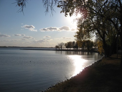 A serene lakeside view with sunlight reflecting on the water, surrounded by trees and a cloudy sky.