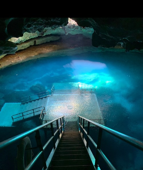 A staircase leads down to a serene, illuminated underground pool surrounded by dark rock formations.