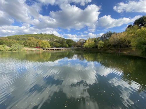 A serene lake reflecting fluffy clouds and trees, surrounded by lush greenery and a distant hill under a blue sky.
