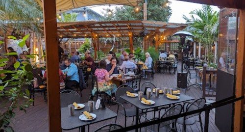 Outdoor dining area filled with people at tables, surrounded by greenery and string lights. Cozy atmosphere.