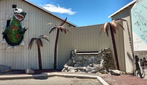 A dinosaur sculpture bursts through a wooden wall, surrounded by metal palm trees and a stone feature.