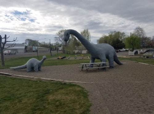 Two large dinosaur sculptures in a park, one towering over a smaller one, with a picnic table nearby and cloudy skies.
