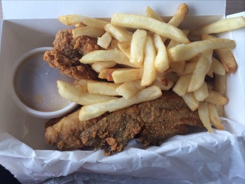 A takeout box containing two pieces of fried fish, a small cup of sauce, and a serving of golden French fries.