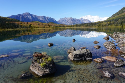 A serene lake reflects mountains and clear blue skies, with rocks and greenery in the foreground.