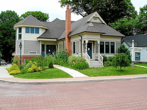 A charming two-story house with a unique roof, surrounded by greenery and a brick-paved street.