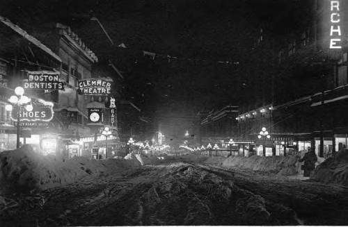 A snowy street at night, lined with vintage storefronts and illuminated signs, creating a nostalgic winter scene.