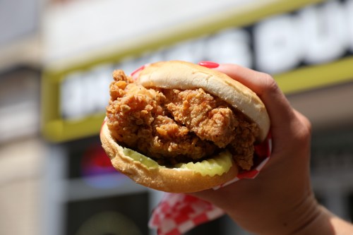 A hand holding a crispy fried chicken sandwich with pickles, set against a blurred background.