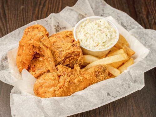 A basket of fried chicken pieces with French fries and a side of coleslaw on a wooden table.