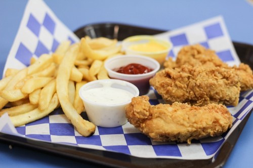 A tray with crispy fried chicken strips, French fries, and dipping sauces in small cups on a blue checkered paper.