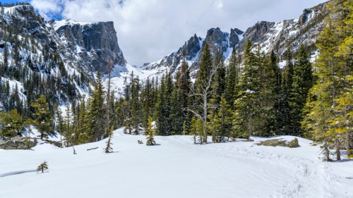 Snow-covered landscape with towering mountains and evergreen trees under a partly cloudy sky.