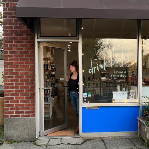 A woman stands at the entrance of a cozy book and bottle shop with large windows and a welcoming atmosphere.