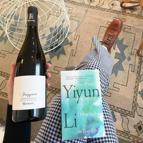 A hand holding a bottle of wine next to a book titled "Dear Friend, Yiyun Li" on a patterned rug.
