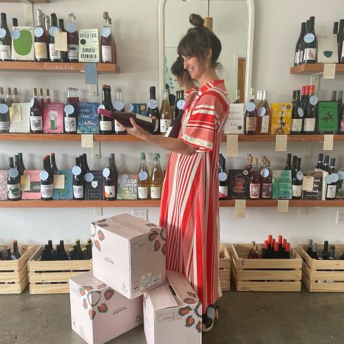 A woman in a striped dress stands in a store, holding a bottle, surrounded by shelves of various beverages and boxes.