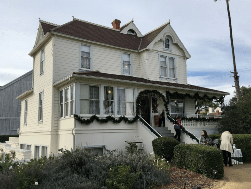 A charming two-story house decorated with greenery, featuring a staircase and people walking outside.