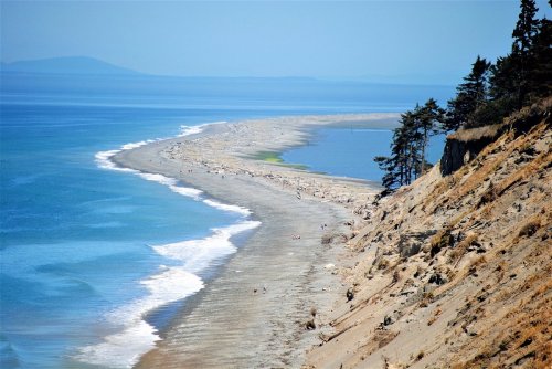 A scenic view of a sandy beach curving along the coastline, with calm blue waters and distant mountains in the background.