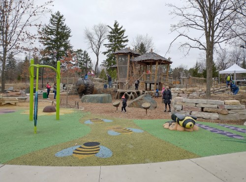 A playground with a wooden structure, climbing rocks, and colorful ground designs, surrounded by trees and children playing.