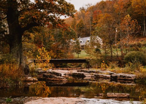A serene landscape featuring a bridge over a stream, surrounded by autumn foliage and a distant house.