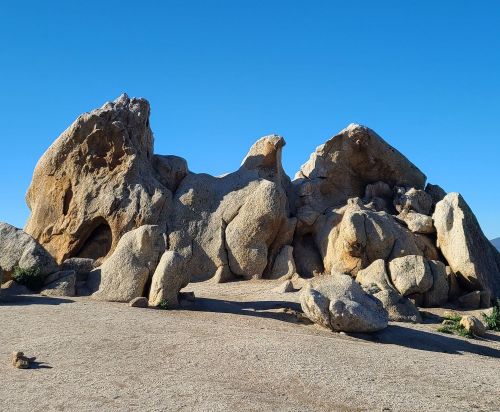 A cluster of large, rugged boulders against a clear blue sky, with a sandy ground and sparse vegetation.