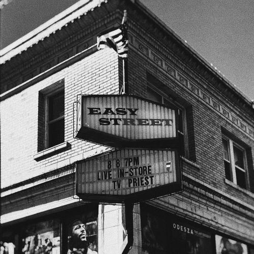 Black and white photo of a storefront sign reading "Easy Street" with event details for TV Priest.