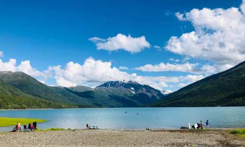 A serene lake surrounded by mountains, with people enjoying the beach and clear blue skies dotted with clouds.