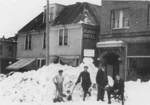 Black and white photo of men standing in front of the Elman Hotel, surrounded by large snowdrifts.