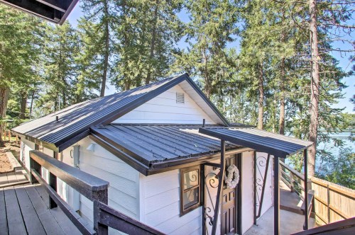 A charming white cottage with a black metal roof, surrounded by tall trees and near a body of water.