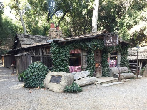 A rustic tavern covered in ivy, surrounded by trees and gravel pathways, with a sign reading "Gold Spring Tavern."