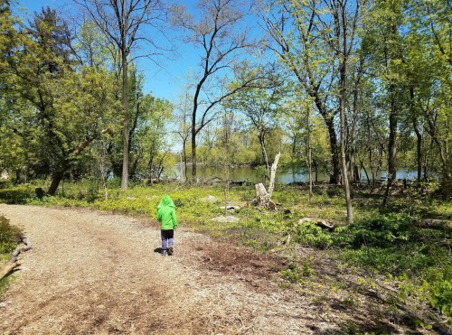 A child in a green jacket walks along a wooded path by a calm lake, surrounded by trees and greenery.