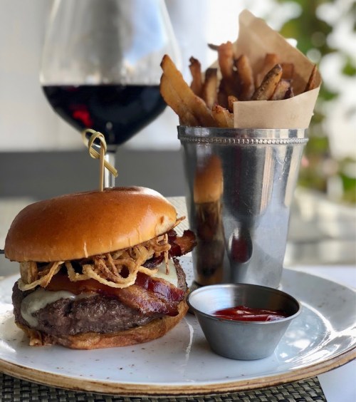 A gourmet burger with bacon, cheese, and crispy onions, served with fries and a side of ketchup, alongside a glass of red wine.