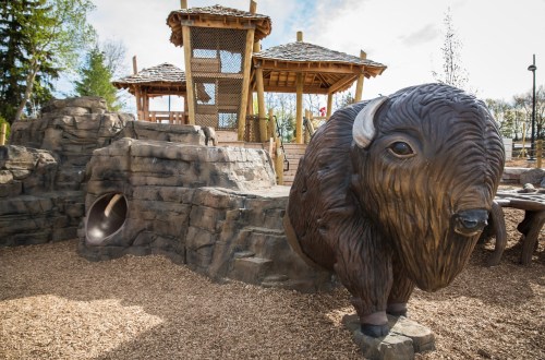 A playground featuring a wooden structure and a large bison sculpture among rocky terrain and wood chips.