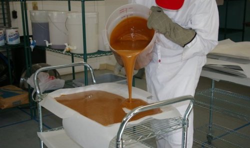A person in protective gear pours a thick, brown liquid from a container onto a flat surface in a lab setting.