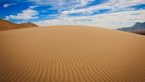 A vast sandy dune with rippled texture under a blue sky with scattered clouds and distant mountains.