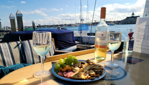 A scenic waterfront table with two glasses of wine, a plate of snacks, and a bottle, overlooking boats and the water.