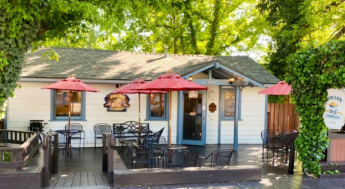 A charming pie shop with red umbrellas, outdoor seating, and greenery surrounding a cozy white building.