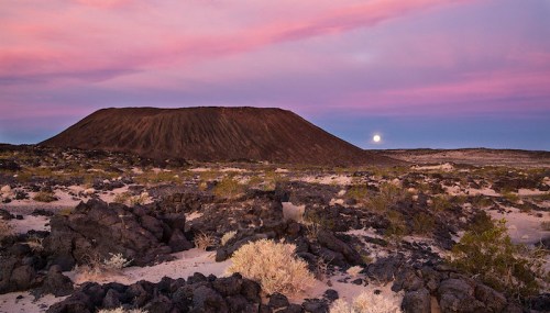 A volcanic landscape at sunset, featuring a dark volcanic cone and a rising moon against a colorful sky.