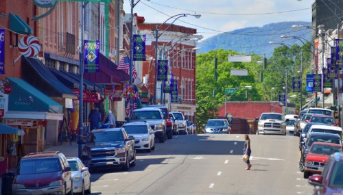 A vibrant street scene with shops, parked cars, and a person walking, set against a backdrop of green hills.