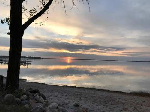 A serene lake at sunset, with reflections on the water and a tree silhouette in the foreground.