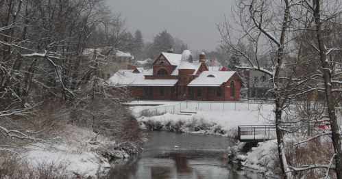 A snowy landscape featuring a red brick building by a river, surrounded by trees and a wintery scene.