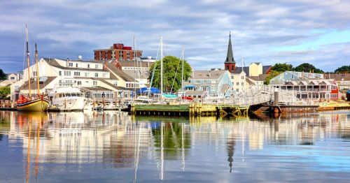 A serene harbor scene with boats, colorful buildings, and a church steeple reflected in calm water under a cloudy sky.
