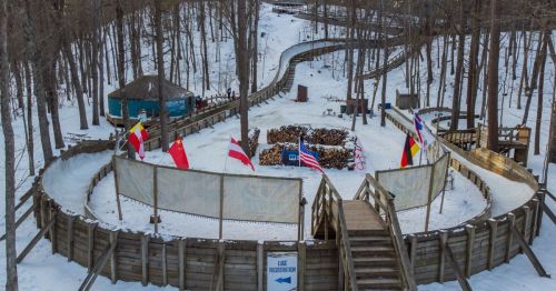 A snowy outdoor ice rink surrounded by trees, with flags and a registration area in the background.