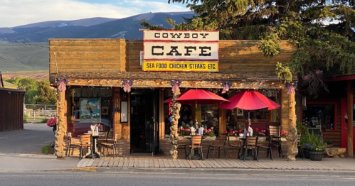 A rustic café with a wooden exterior, red umbrellas, and outdoor seating, set against a mountain backdrop.