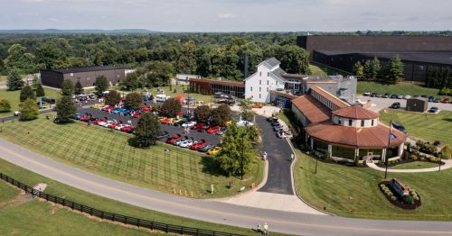 Aerial view of a car show with colorful vehicles parked outside a modern building, surrounded by green trees and fields.