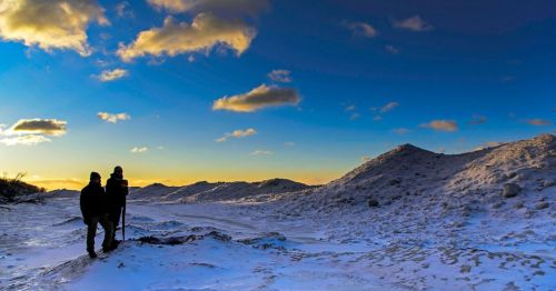 Two hikers silhouetted against a vibrant sunset over a snowy landscape with rolling hills and a clear blue sky.