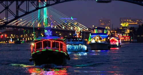 Colorful boats adorned with lights navigate a river at night, with a bridge and city skyline in the background.